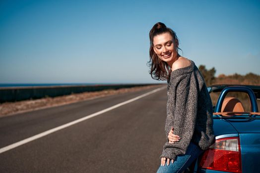 Young woman near roofless car and smiling outdoors on sunny day near the empty road. No people, sportive blue car