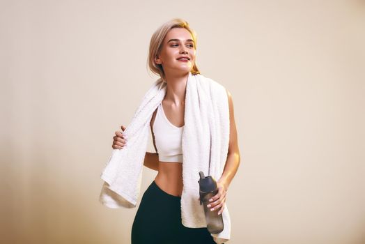 Tired but happy Cute and young sporty woman in sports clothing with towel on shoulders looking aside and smiling while standing in studio. Sport. Active lifestyle