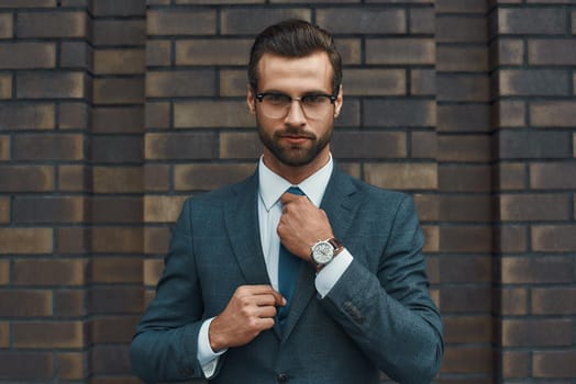 Perfect look. Confident businessman in full suit adjusting his tie and looking at camera while standing against brick wall. Business concept. Stylish people