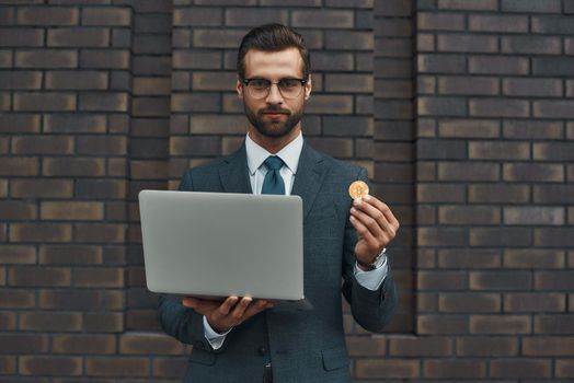 Our future. Handsome businessman in formal wear and eyeglasses holding laptop and bitcoin in another hand while standing against brick wall. Business concept. Cryptocurrency