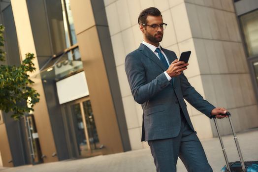 Ready to help everywhere. Young and handsome bearded man in suit carrying suitcase and looking at his smartphone while standing outdoors. Travelling. Business concept