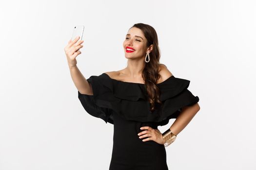 Beautiful woman in black dress taking selfie on party, standing over white background with smartphone.