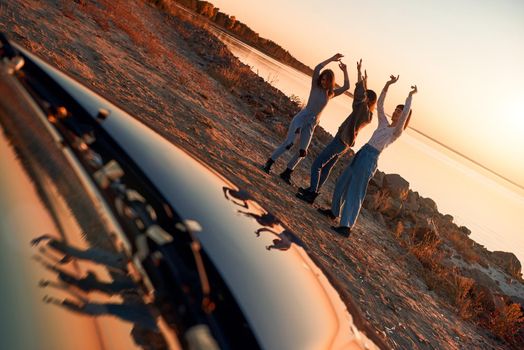 Cheerful young three women are dancing near cabriolet. Beautiful sunset at the background