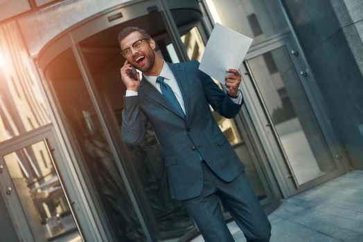 Great news Portrait of happy bearded businessman in full suit talking by phone with client and smiling while walking outdoors. Business concept