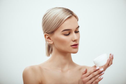 Care about your skin. Portrait of attractive young blonde woman holding face cream while standing against grey background. Skin care. Beauty concept. Cosmetics concept