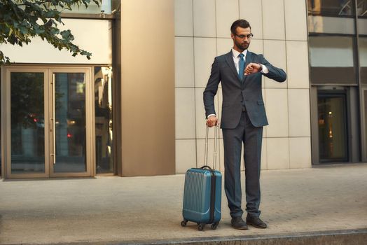 I am in a hurry. Full length of young and handsome bearded man in suit carrying suitcase and looking at watch on his hand while standing outdoors. Travelling. Business trip