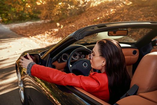 Beautiful girl in the roofless car. Autumn season, red coat and jeans, stylish sunglasses on head. Close-up, rear view