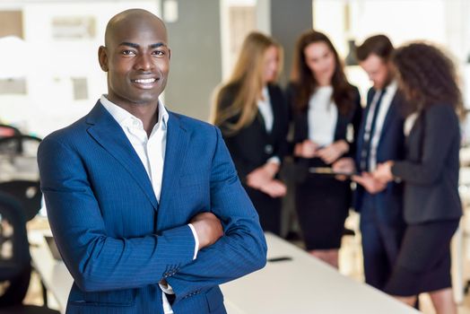 Black businessman leader looking at camera in modern office with multi-ethnic businesspeople working at the background. Teamwork concept. African man wearing blue suit.