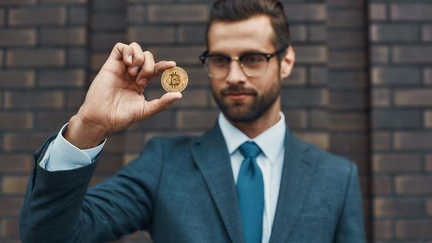 Digital currency. Handsome businessman in formal wear and eyeglasses holding bitcoin in one hand and looking at it while standing against brick wall. Business concept. Cryptocurrency