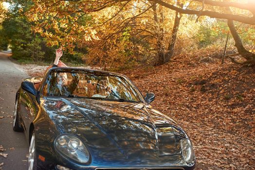 A black roofless car driving fast on the countryside asphalt road against morning sky with a beautiful sunrise. Autumn season, falling leaves