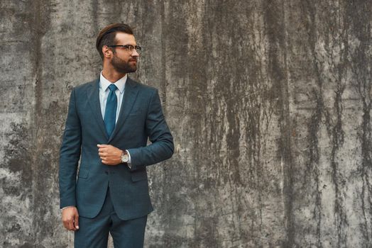 Great style. Confident bearded businessman in full suit looking away while standing against grey stone wall outdoors. Business concept. Stylish people