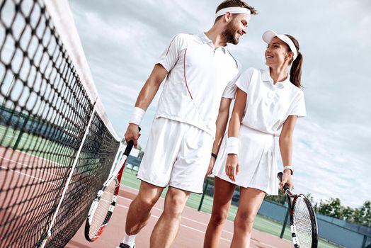 Beautiful woman and handsome man in white sportswear with rackets in their hands are smiling to each other while talk on the court