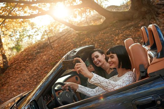 Side view of two women friends in cabliolet. They are laugthing and making selfie. Happy journey