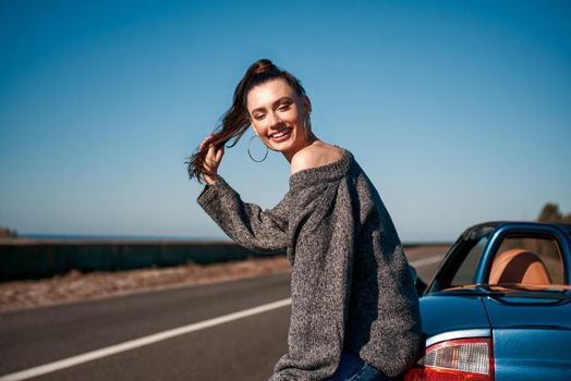 Young woman near roofless car and smiling outdoors on sunny day near the empty road. No people, sportive blue car