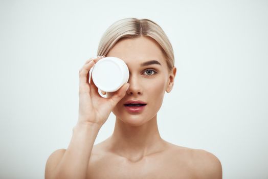 Deep hydration. Portrait of cute young blonde woman showing face cream while standing against grey background. Skin care. Beauty concept. Cosmetics concept