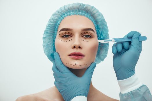 Face lift surgery. Portrait of beautiful young woman in blue medical hat having cosmetic face surgery. Plastic surgeon in blue gloves holding scalpel. Beauty concept