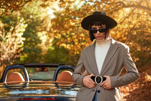 Attractive young woman posing near cabriolet. She wears black hat and sunglasses. Stylish autumn.