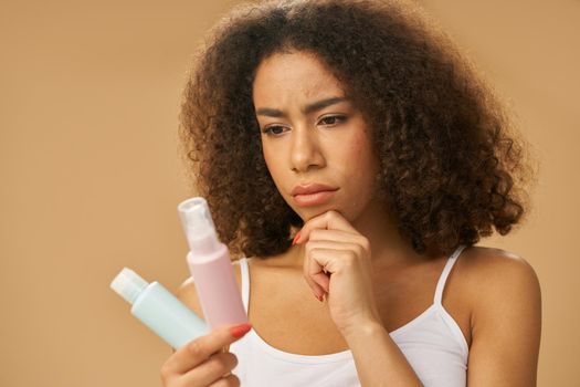 Attractive young woman with curly hair looking doubtful while choosing between two beauty products, posing isolated over beige background. Skincare concept