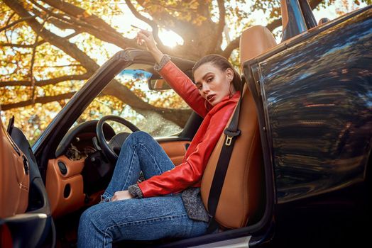 Beautiful girl in the roofless car. Autumn season, red coat and jeans, stylish sunglasses on head. Skin salon