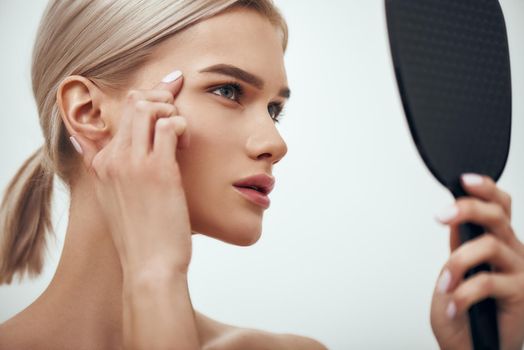 Mimic wrinkles. Profile portrait of young attractive woman touching her face and looking in small mirror while standing against grey background. Skin care. Beauty concept