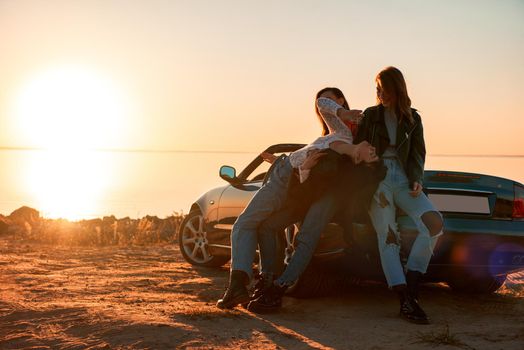 Cheerful young three women standing near cabriolet. Beautiful sunset at the background