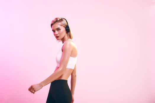 Time for music. Side view of sporty blonde woman in headphones listening music while standing against pink background. Sport. Music concept