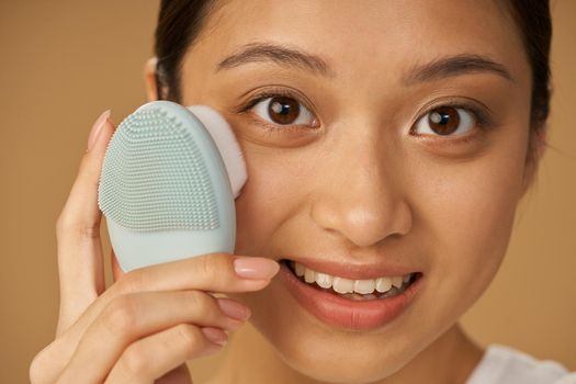 Face closeup of adorable young woman smiling at camera while using silicone facial cleansing brush, posing isolated over beige background. Skincare concept