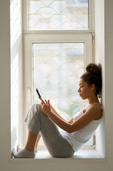 Side view of relaxed young woman reading a book using tablet pc while sitting by the window at home. Lifestyle, leisure concept. Side view