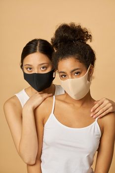 Portrait of two beautiful young diverse women wearing protective facial masks looking at camera while posing together isolated over beige background. Safety, pandemic concept