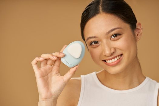 Adorable young woman smiling at camera, holding silicone facial cleansing brush, posing isolated over beige background. Washing accessory. Skincare concept