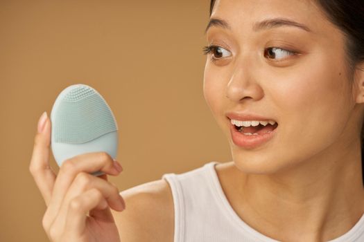 Close up portrait of cute young woman looking surprised while holding silicone facial cleansing brush, posing isolated over beige background. Washing accessory. Skincare concept