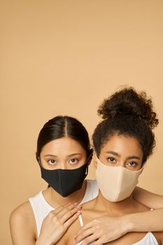 Portrait of two adorable young diverse women wearing protective facial masks looking at camera while posing together isolated over beige background. Safety, pandemic concept