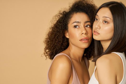 Portrait of two gorgeous mixed race young women with perfect skin looking away while posing isolated over beige background. Natural beauty, skincare, diversity concept