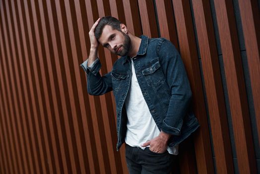 Freestyle. Young man standing leaning on wooden wall isolated touching hair looking aside pensive