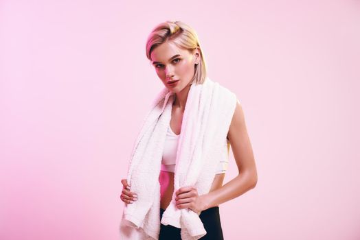 Time for rest. Attractive young blonde woman with towel on shoulders looking at camera while standing against pink background in studio. Sport. Active lifestyle. Workout