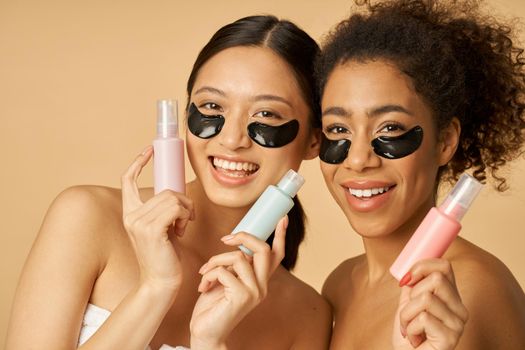 Two lovely young women with hydro gel under eye patches smiling at camera, holding bottles with beauty product, posing isolated over beige background. Skincare concept