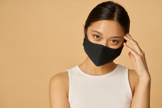 Confident young woman wearing black facial mask looking at camera, posing isolated over beige background. Safety, pandemic concept