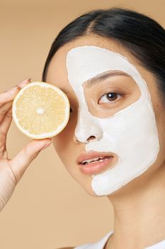 Beauty portrait of young woman with facial mask applied on half of her face holding a slice of lemon, posing isolated over beige background. Skincare, natural cosmetics concept. Vertical shot