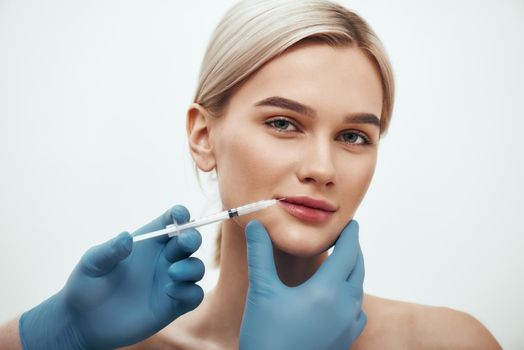 Cosmetic injection. Young beautiful woman looking at camera and smiling while doctors hand making an injection in her lips. Studio shot. Beauty concept