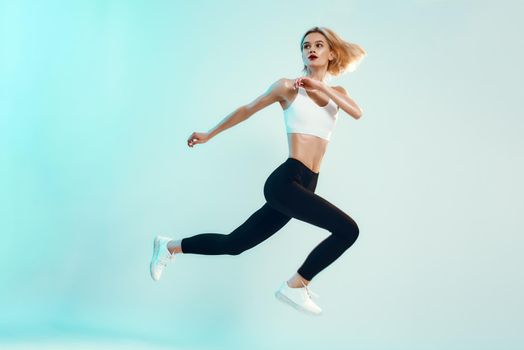 No limits Sporty and young woman with perfect body in white top and black leggings jumping against blue background in studio. Sport. Active lifestyle. Studio shot
