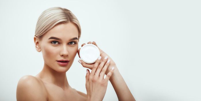 You need a special face cream. Cute young blonde woman showing face cream while standing against grey background. Skin care. Horizontal web banner. Cosmetics concept