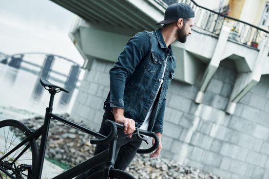 Outdoors leisure. Young stylish man walking on city street with bicycle looking aside pensive