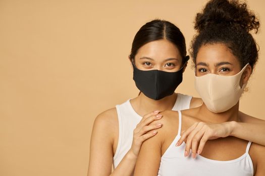 Portrait of two beautiful young diverse women wearing protective facial masks looking at camera while posing together isolated over beige background. Safety, pandemic concept