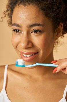 Close up portrait of natural young mixed race woman brushing teeth, posing isolated over beige background. Dental health care concept