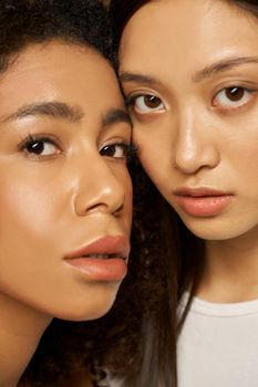 Face closeup of two beautiful mixed race young women with perfect glowing skin looking at camera while posing together. Skincare, diversity concept. Selective focus. Vertical shot