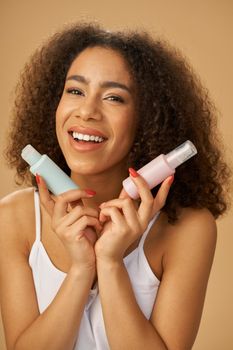 Happy young mixed race woman looking excited, holding two bottles with beauty product while posing isolated over beige background. Skincare concept. Vertical shot