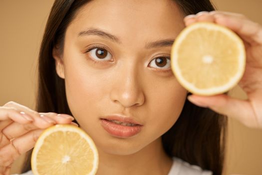 Close up portrait of charming mixed race young woman looking at camera, holding lemon cut in half while posing isolated over beige background. Health and beauty concept