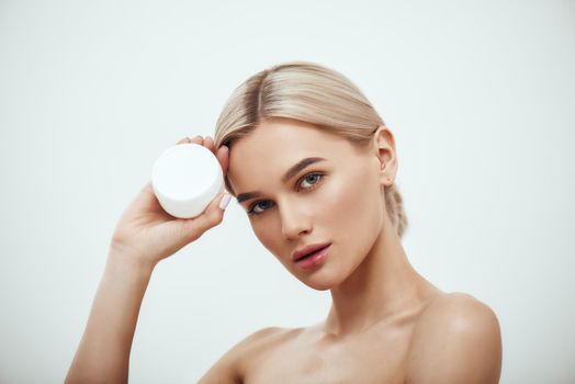 Best Facial Cream. Portrait of gorgeous young blonde woman showing face cream while standing against grey background. Skin care. Beauty concept. Cosmetics concept