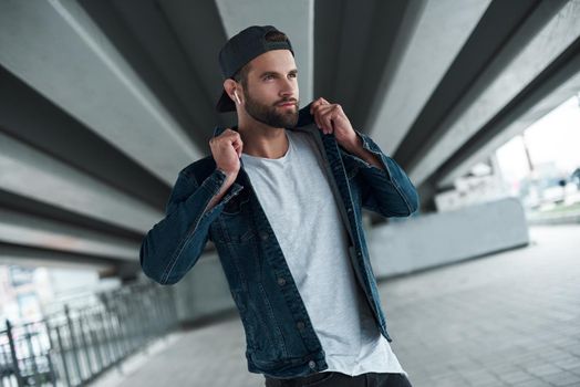 Outdoors leisure. Young stylish man walking on city street wearing wireless earphones listening to music looking aside smiling joyful