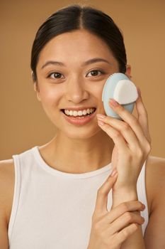 Excited young woman smiling at camera while using silicone facial cleansing brush, posing isolated over beige background. Skincare concept